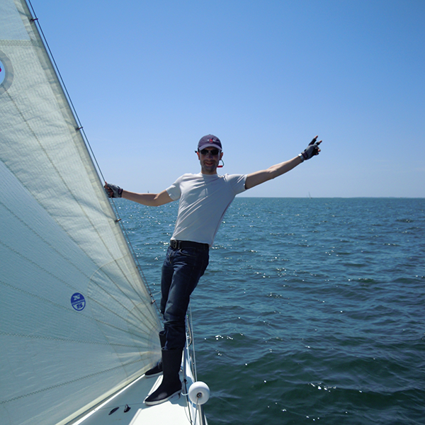 Prise de vue de la proue d’un voilier, depuis le cockpit, avec moi au premier plan, tenant le guindant du génois de la main droite et faisant de la gauche un signe index et majeur tendus comme des ciseaux.