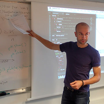 Cliché de face, en salle de cours, un tableau blanc bardé de formules mathématiques et un écran de projection avec des lignes de code en surimpression.